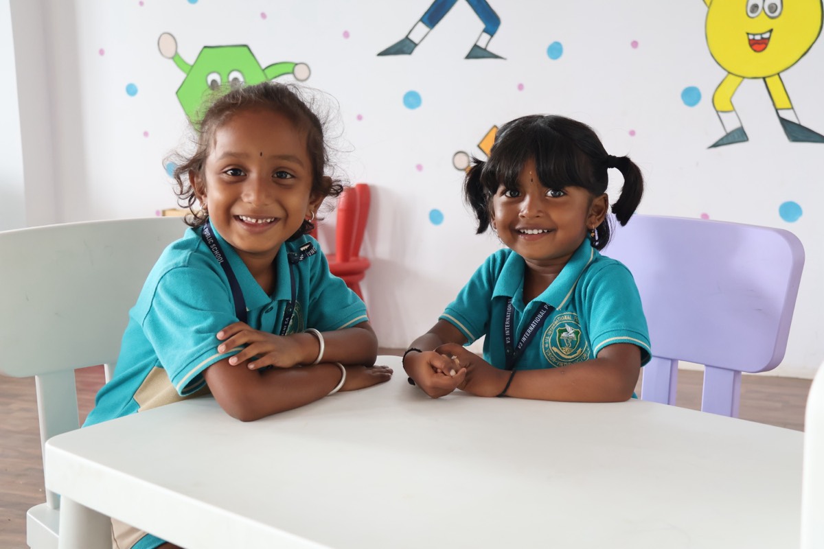 Two V3 students in school uniform smiling together at a classroom table